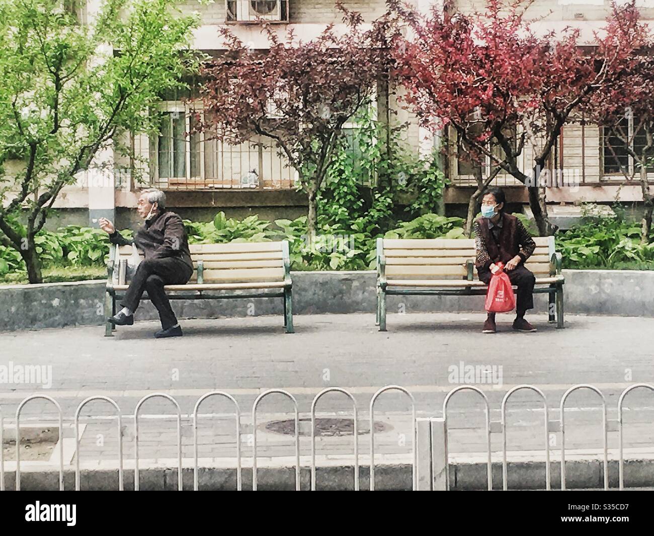 Beijing, China- April 17, 2020: social distancing in China. An old man and old woman sitting on different benches outdoors. - Smartphone Captured Stock Image