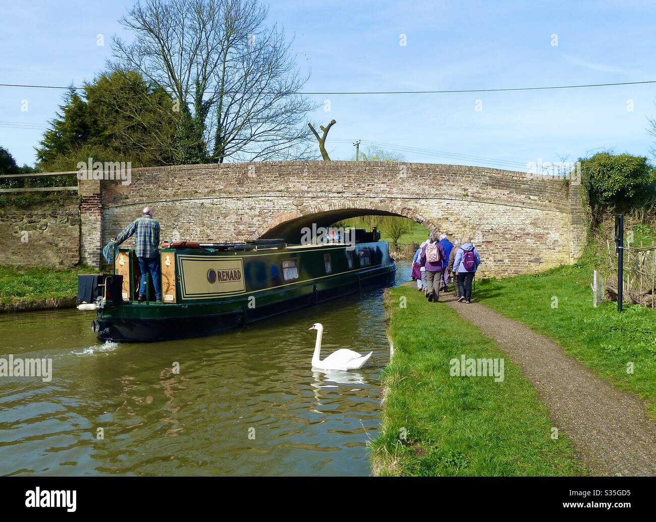 Ramblers walking along the grand Union canal as a narrow passes under a bridge with a swan swimming nearby Stock Photo