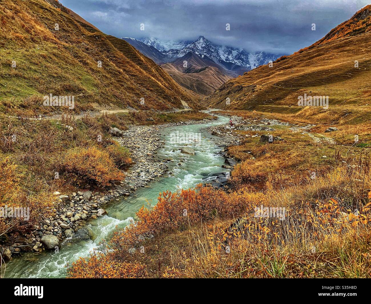 River running through a mountain valley covered in autumnal colours in the shadows of a snowy mountain peak in Ushguli, Georgia. Stock Photo