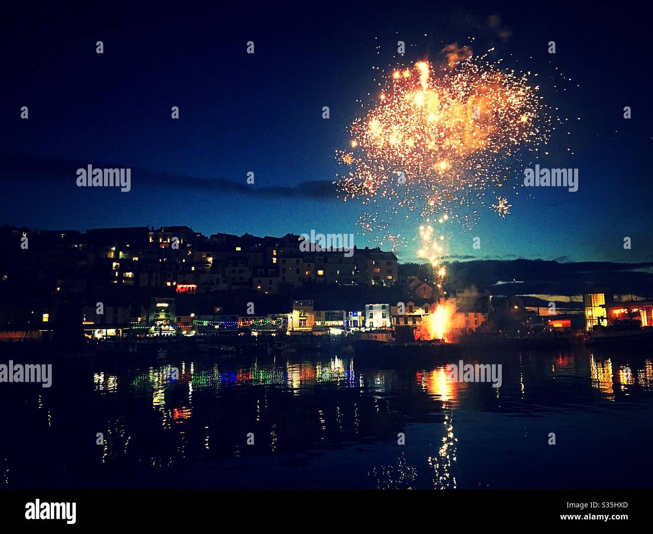 Brixham Harbour: Firework in the night sky over a beautifully lit up harbour side town, which reflects in the water. - Smartphone Captured Stock Image