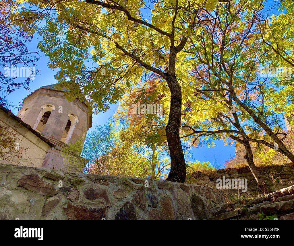 A monastery in Georgia alongside some autumnal coloured trees, glowing in the sun. Against a blue sky. - Smartphone Captured Stock Image
