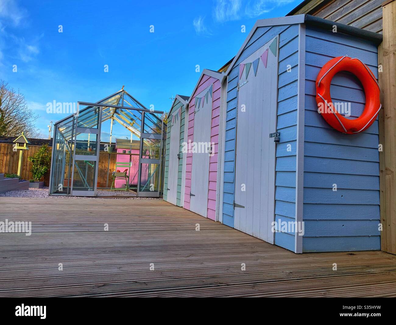 Three colourful beach huts in a garden with a greenhouse, bird table and a deck on a sunny day. - Smartphone Captured Stock Image Three colourful beach huts in a garden with a greenhouse, bird table and a deck on a sunny day. - Smartphone Captured Stock Image