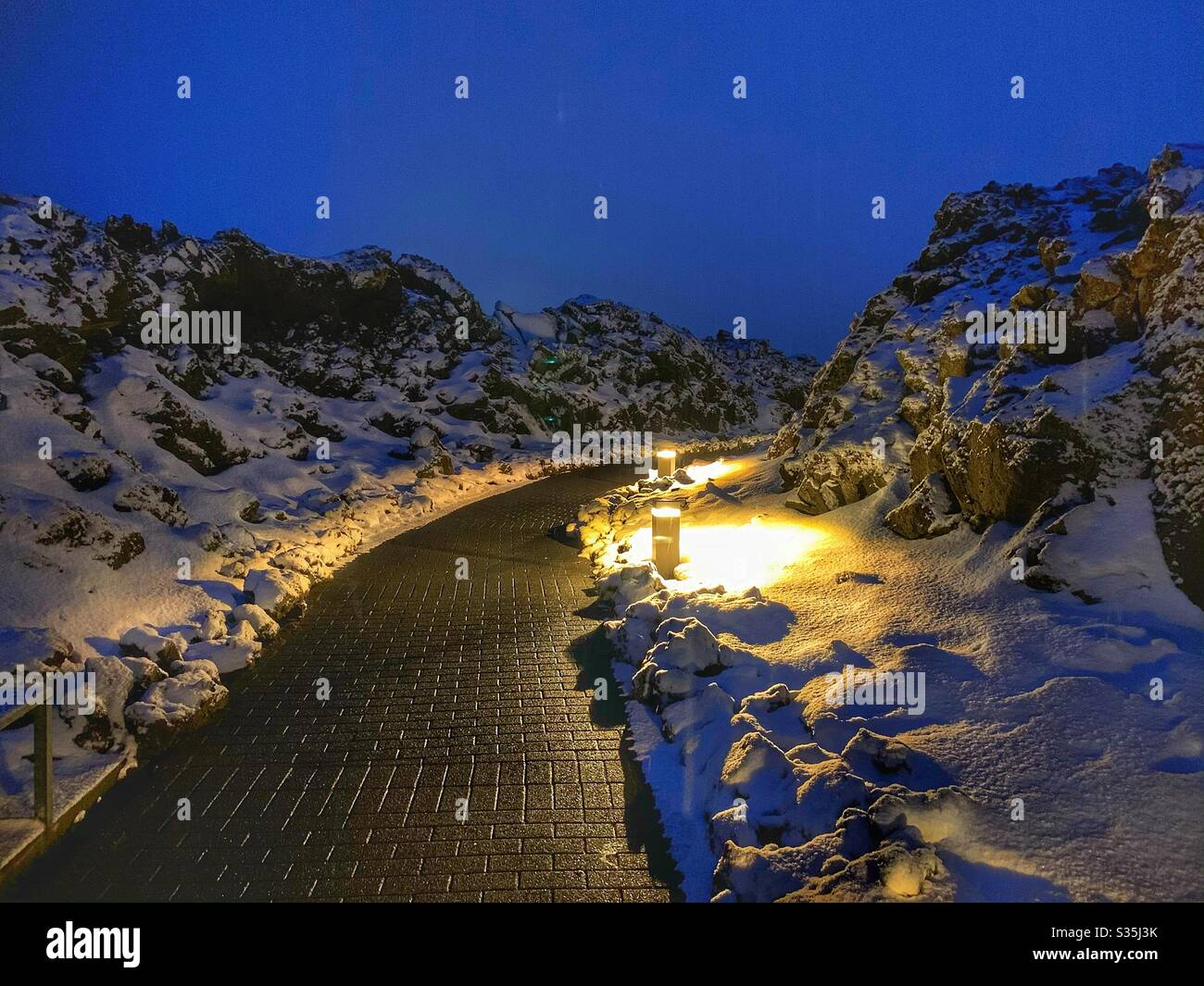 Pathway surrounded by black volcanic rock, covered in snow and lit by walkway lighting at blue hour. Entrance to The Blue Lagoon, Iceland. Stock Photo