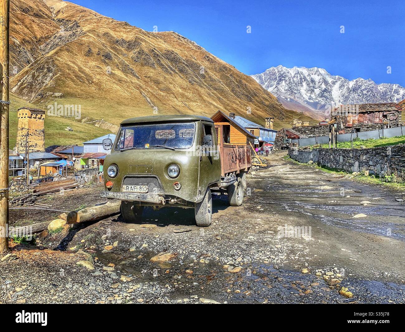 An old soviet truck is parked on the track at the entrance to the UNESCO mountain village of Ushguli, Georgia. Surrounded by snowy mountains and ramshackle houses. - Smartphone Captured Stock Image