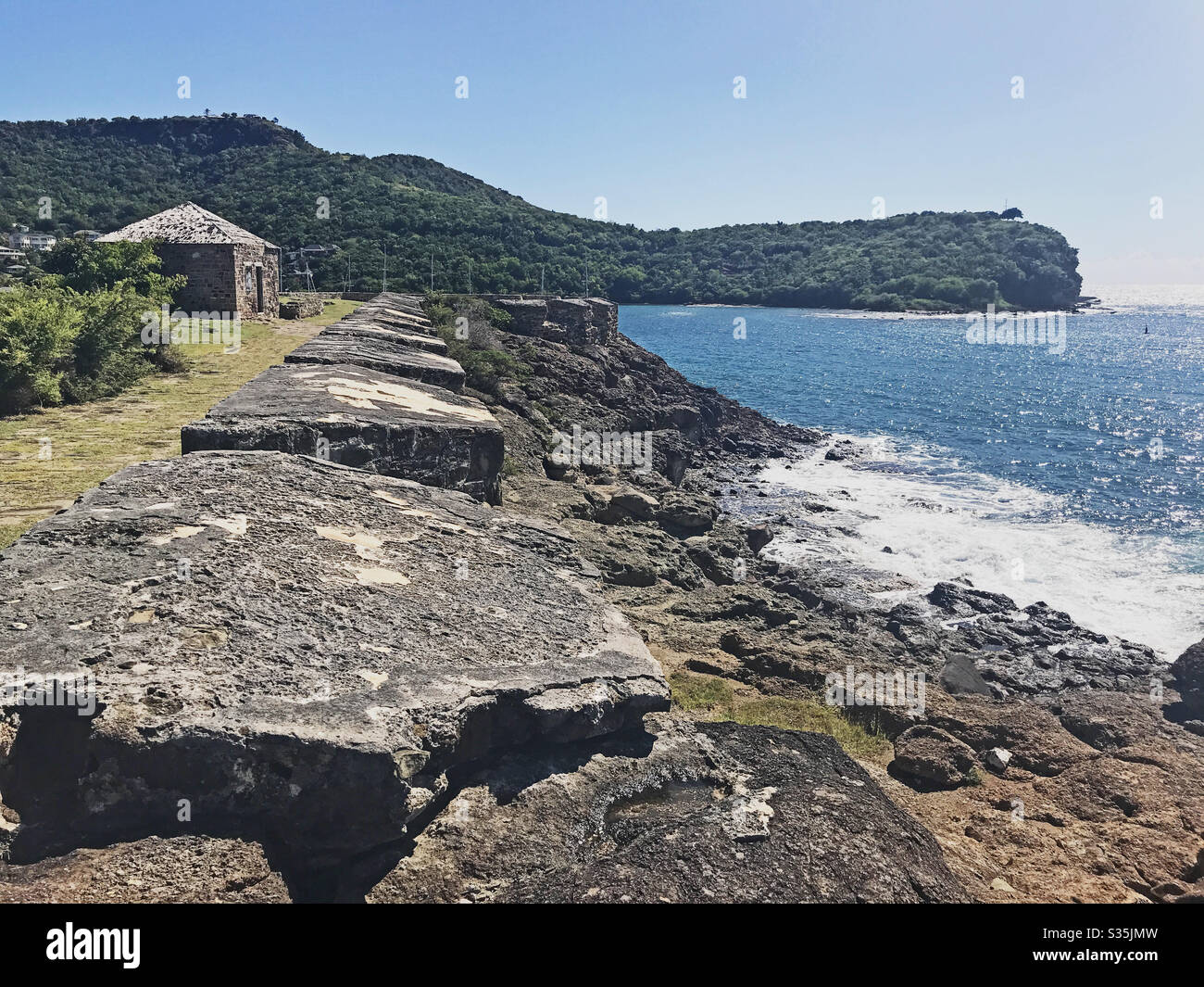 Antigua coastline, Guard House and outer fort wall with cannon ports as seen from the Fort Berkeley Peninsula in Antigua and Barbuda, Caribbean, Lesser Antilles, West Indies with blue sky copy space. - Smartphone Captured Stock Image