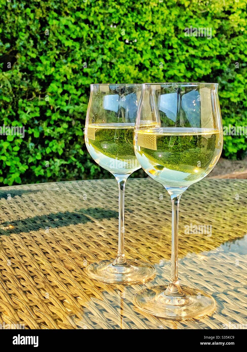 Two glasses of white wine on the glass table top of garden furniture in morning light Stock Photo
