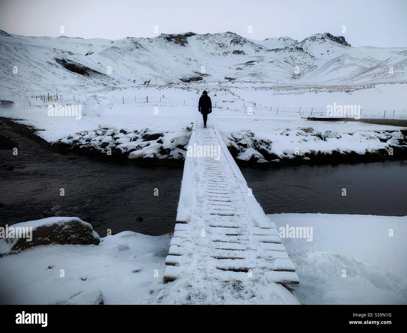 Isolation. A dark figure stands with their back to the camera on the end of a snowy bridge over a river. A snowy mountainous landscape in the background. - Smartphone Captured Stock Image