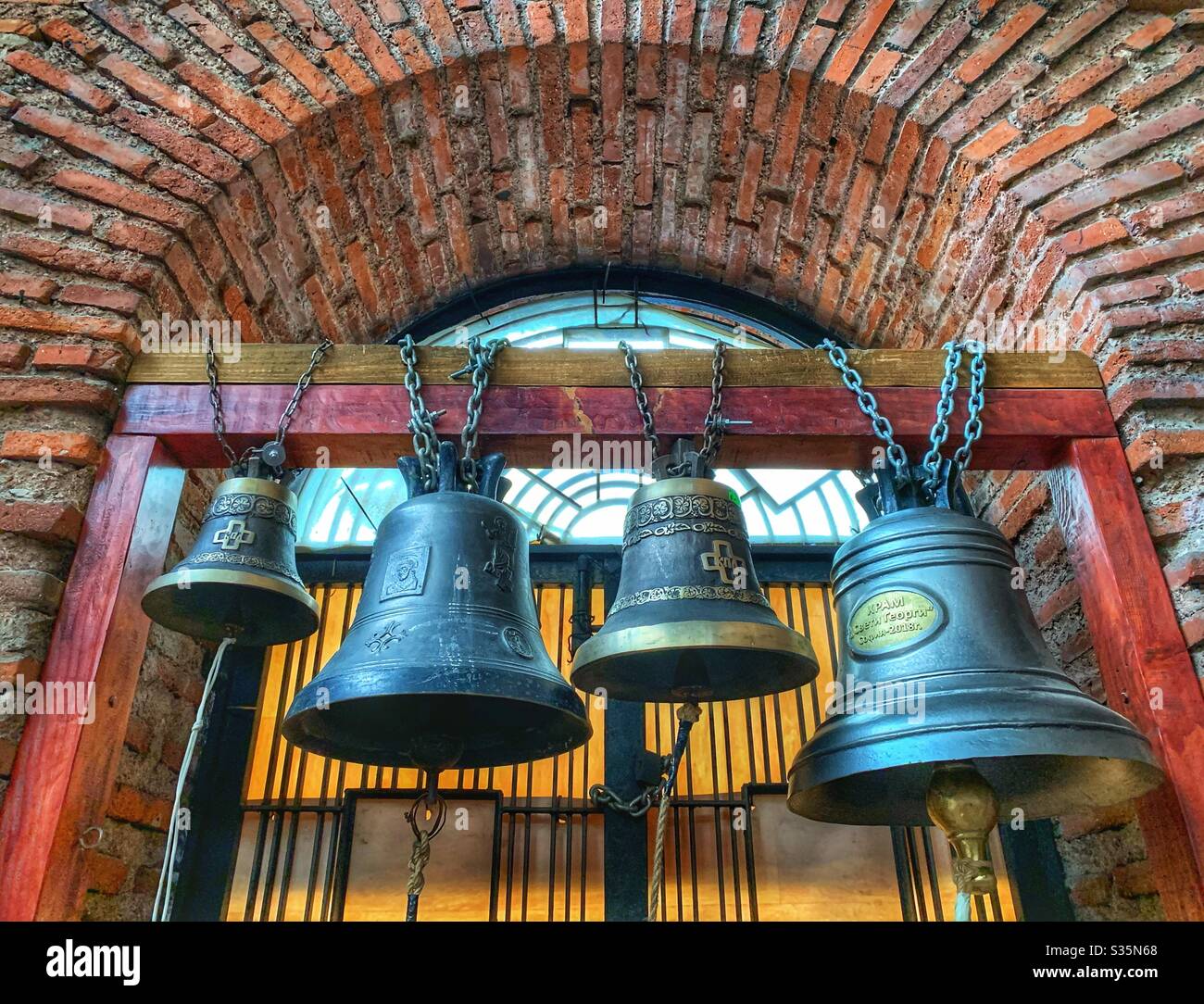 Four bells chained to a wooden support, surrounded by a brick archway. St George Rotunda Church, Bulgaria. - Smartphone Captured Stock Image