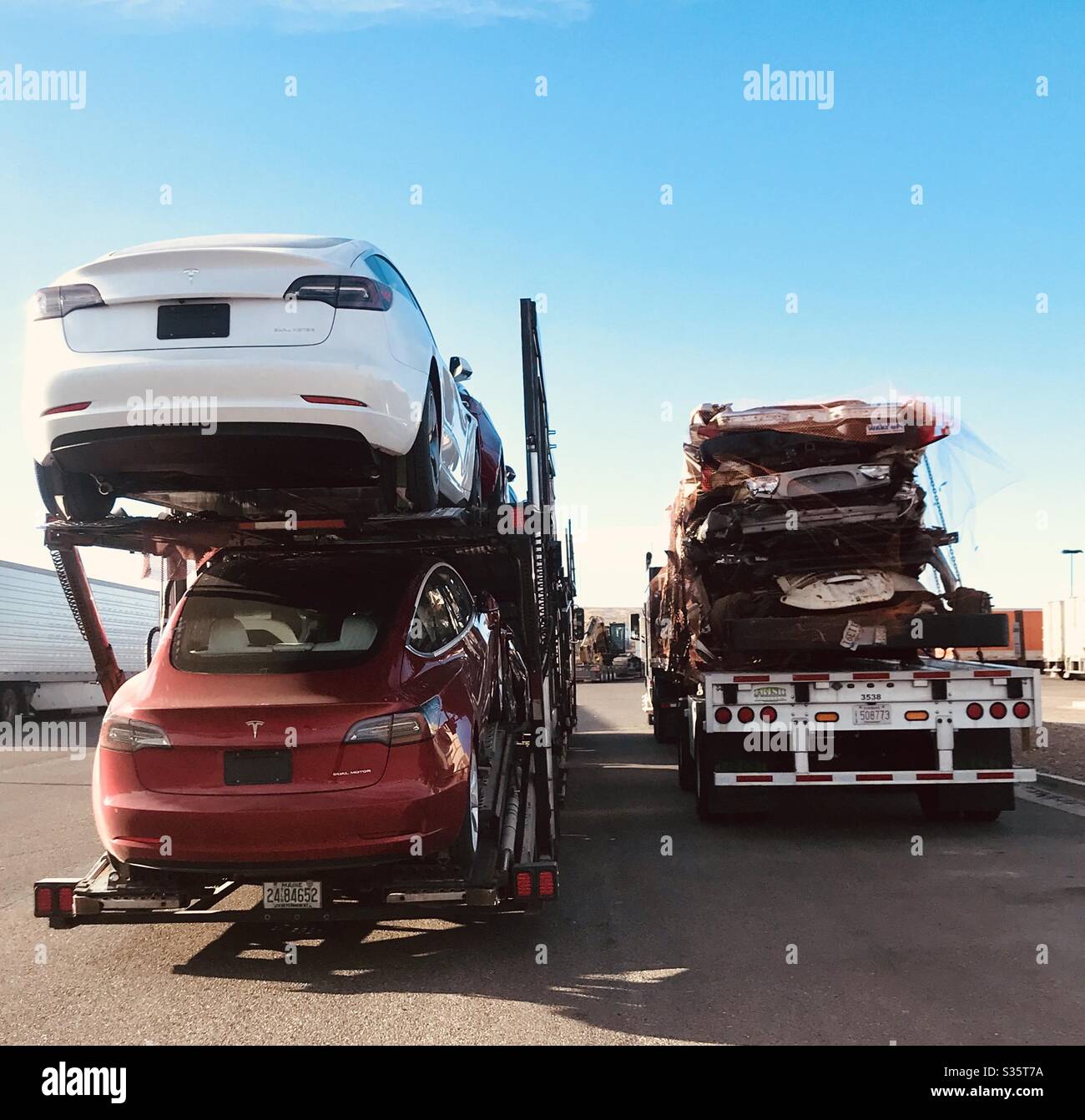 Two semi trucks next to each other, one carrying brand new modern Tesla cars, and the other smashed up old cars. Stock Photo