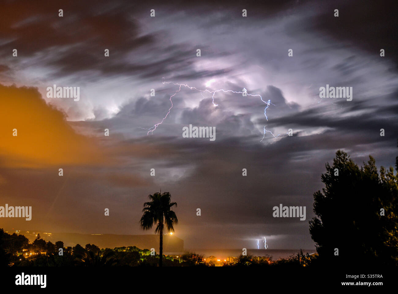 Electric storm out to sea with palm tree and town lights in foreground Stock Photo