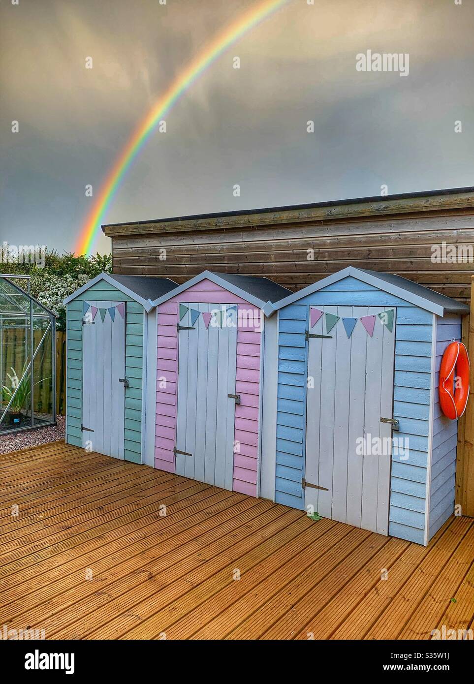 Garden beach huts sat on a deck against a dark sky with a vibrant rainbow. - Smartphone Captured Stock Image