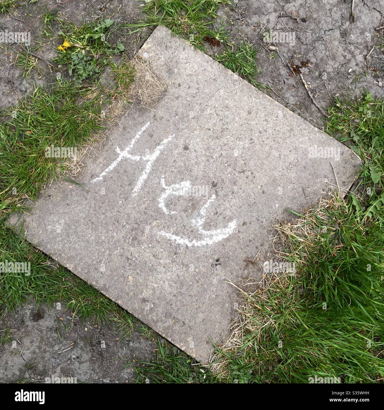 A photo looking down at a paving slab with the word ‘hey’ written on it in white chalk. Paving stone on a rural path with grass clumps. Fun communication in the time of isolation. Stock Photo