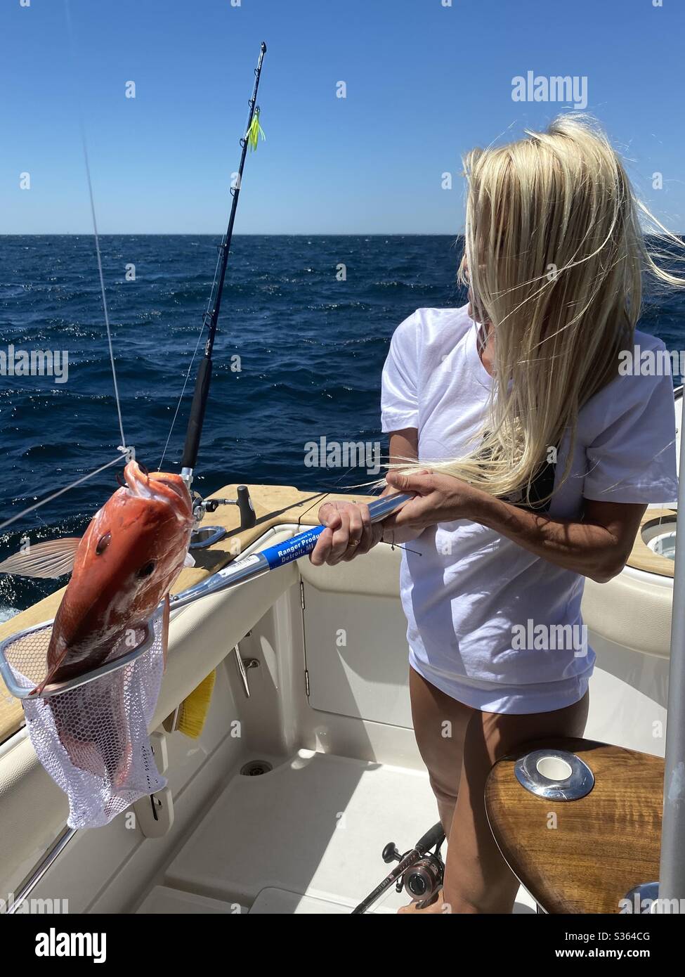 Young woman with long blonde hair on a boat holding a red snapper fish she caught - Smartphone Captured Stock Image
