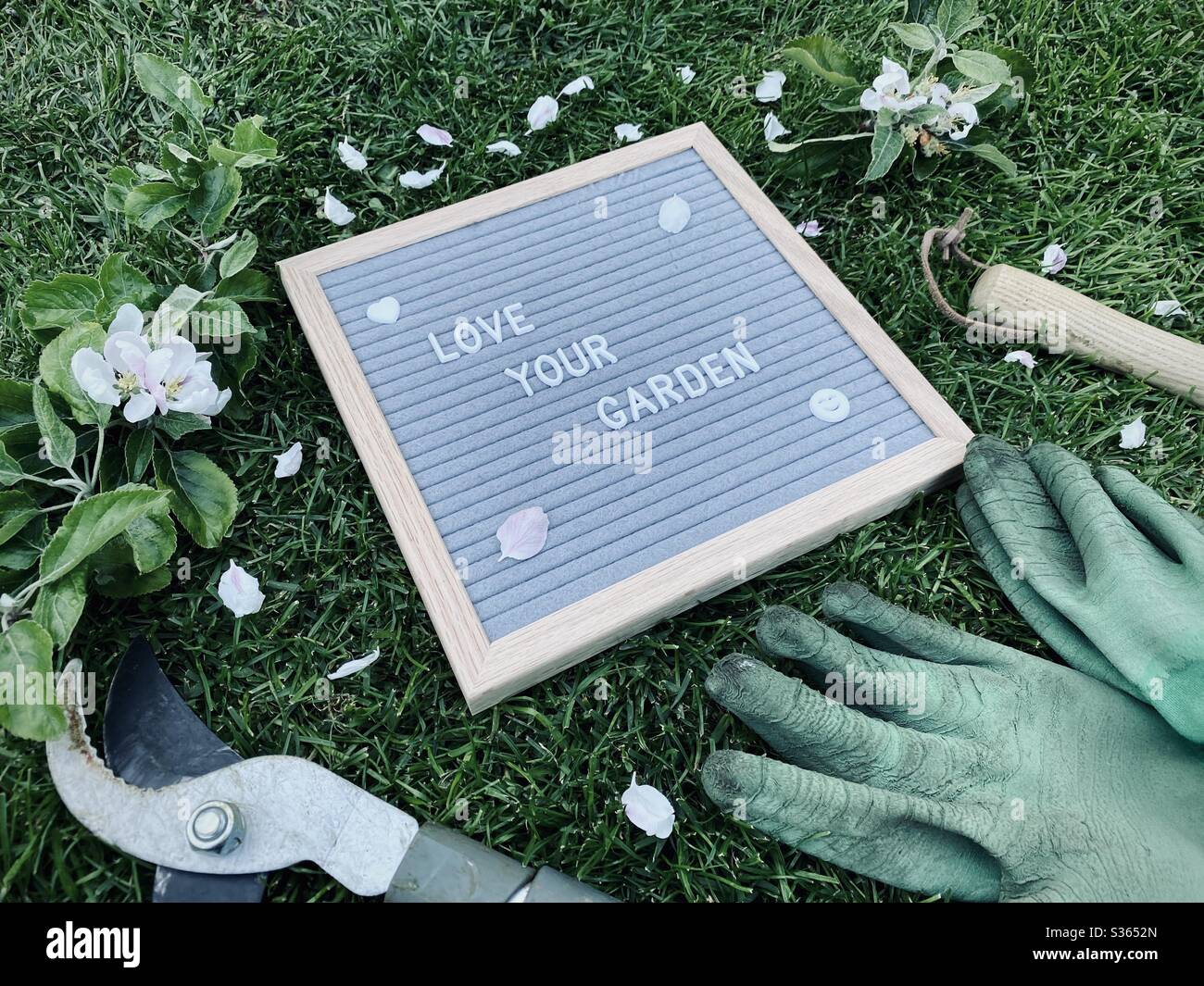 Love your garden. Gardening concept with felt letter board, handheld prune, dirty gloves and a pruned branch with blossom from an apple tree on a green lawn - Smartphone Captured Stock Image