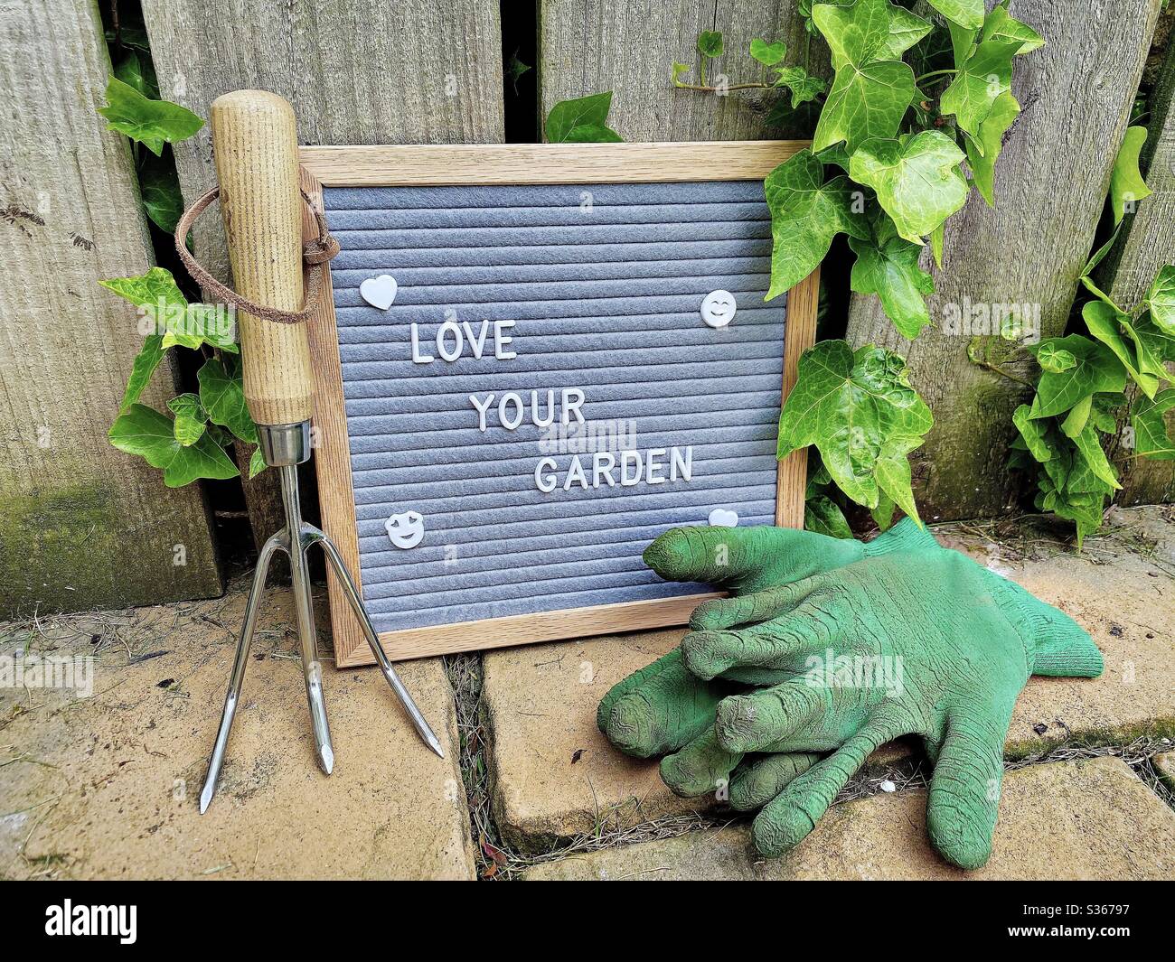 Love your garden. Gardening concept with felt letter board, dirty gloves and a handheld fork on a stone patio against a weathered fence background with overgrown ivy. - Smartphone Captured Stock Image