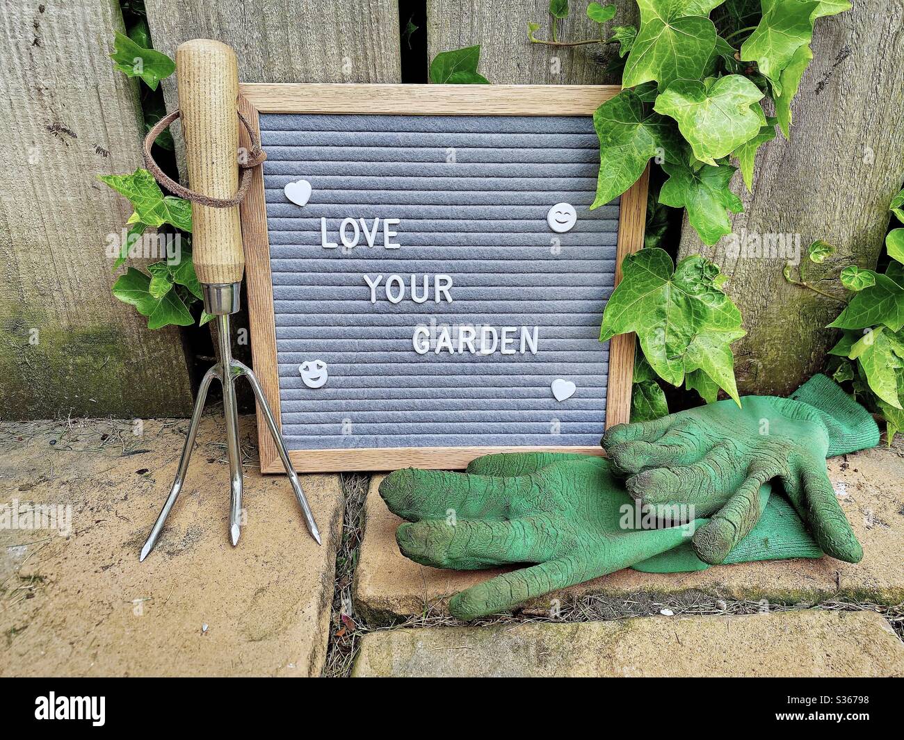 Love your garden. Gardening concept with felt letter board, dirty gloves and a handheld fork on a stone patio against a weathered fence background with overgrown ivy. - Smartphone Captured Stock Image