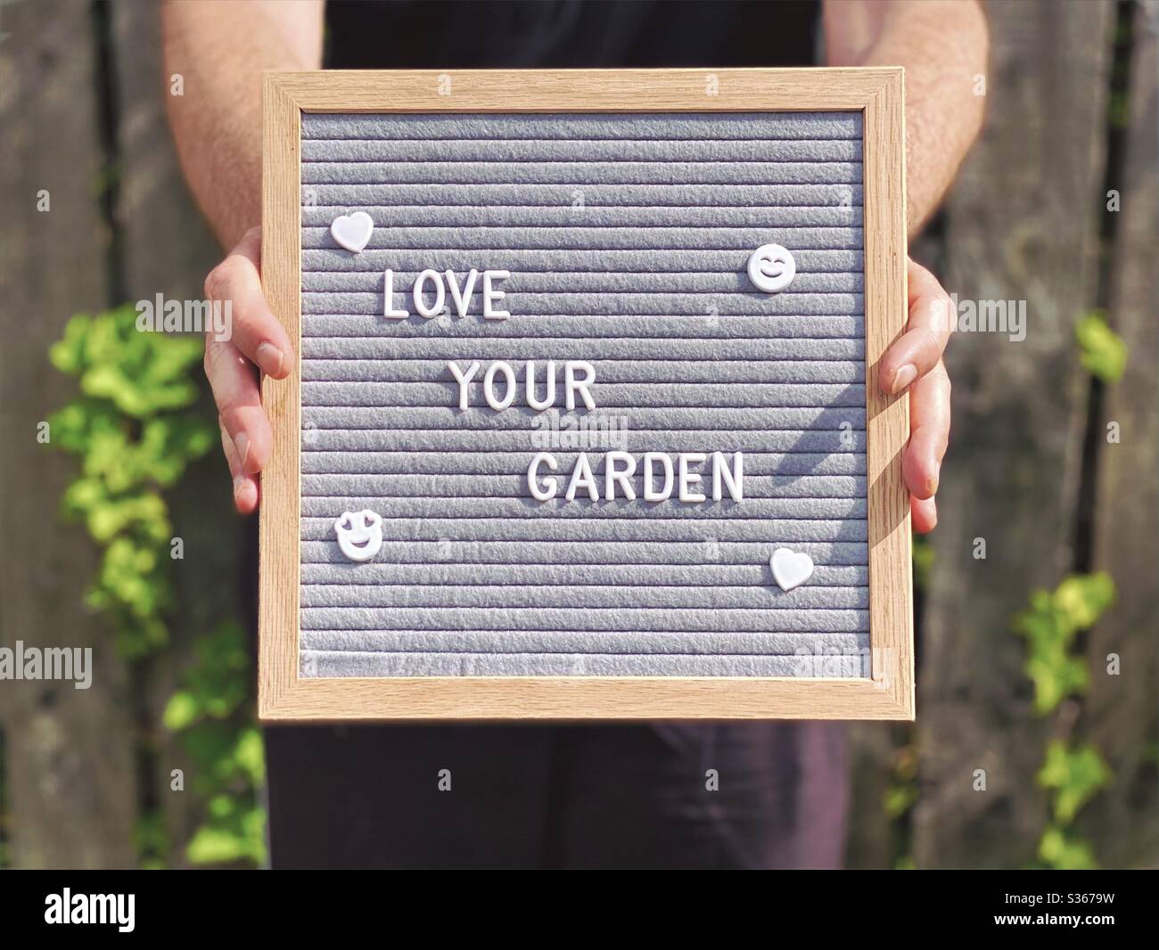 Love your garden. Gardening concept with felt message board in the hands of a Caucasian man standing against a weathered fence with overgrown ivy seen blurry in background. - Smartphone Captured Stock Image