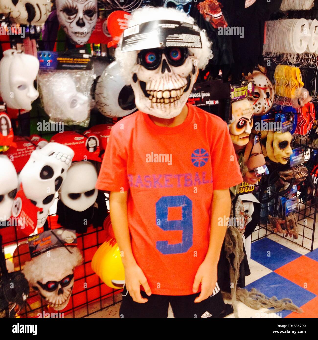 Young boy in a toy store trying on  a scary skeleton Halloween mask by a large display of masks. Stock Photo