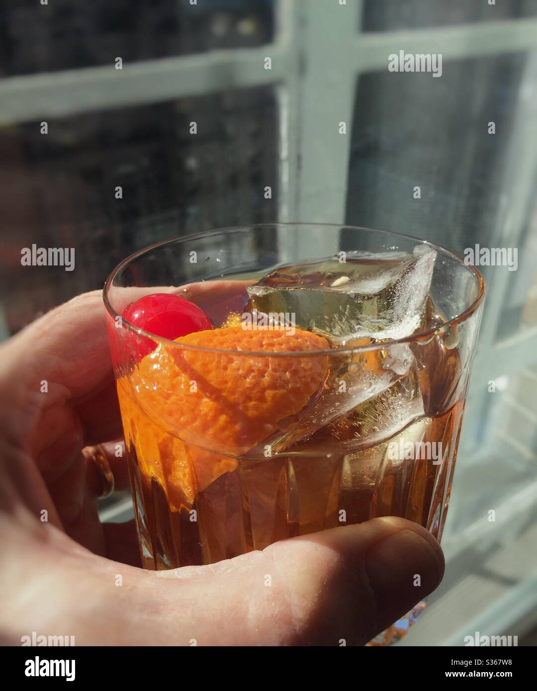 Man holding a Manhattan cocktail in a crystal glass with orange and cherry garnish - Smartphone Captured Stock Image