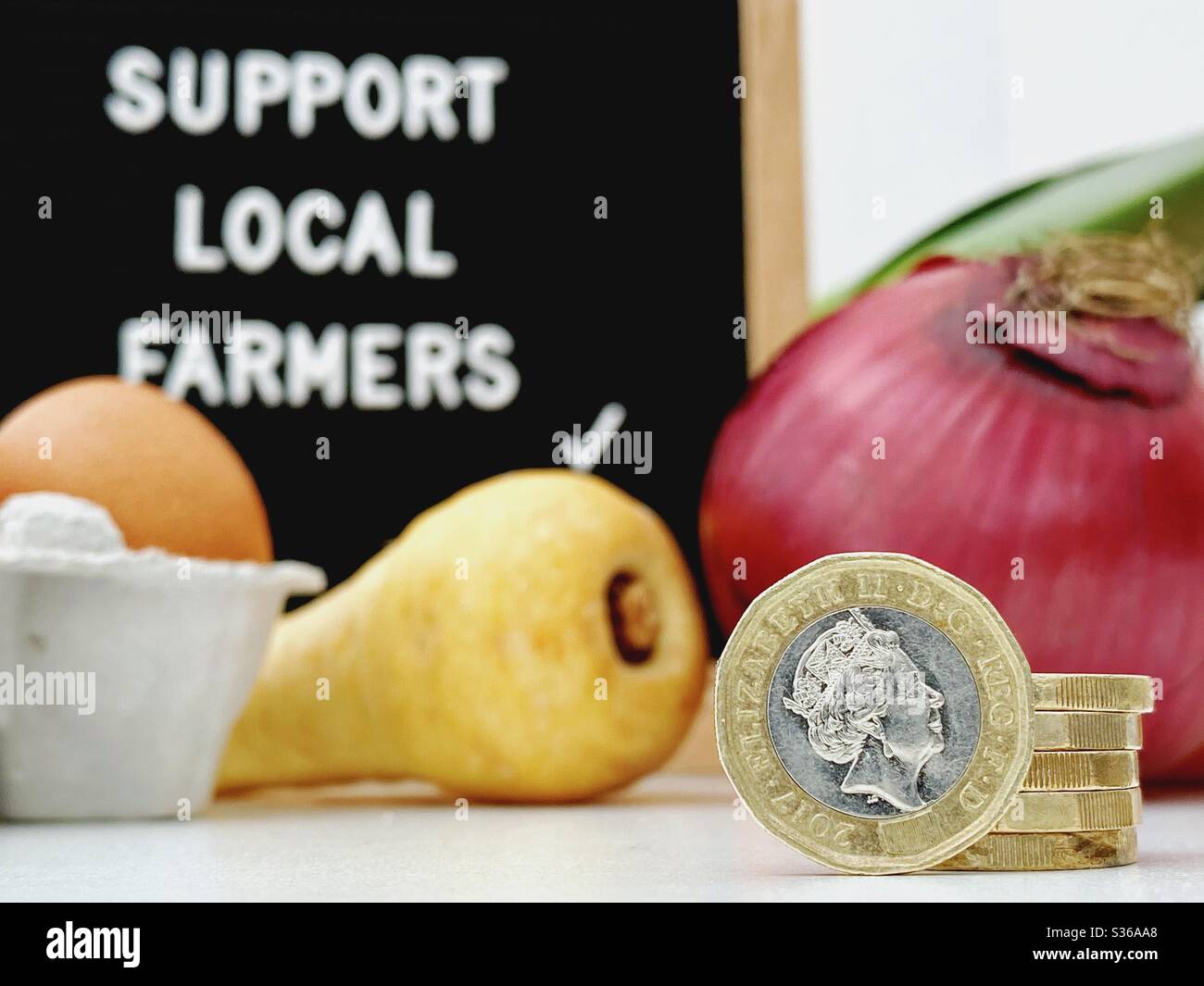 A selective focus view of a blurry farmer’s market sign behind fresh vegetables. Support local farmers. Fresh vegetables with British pound coins and loose change in foreground - Smartphone Captured Stock Image