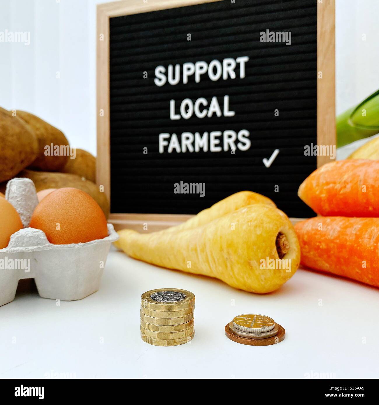 A selective focus view of a blurry farmer’s market sign behind fresh vegetables. Support local farmers. Fresh vegetables with British pound coins and loose change in foreground - Smartphone Captured Stock Image