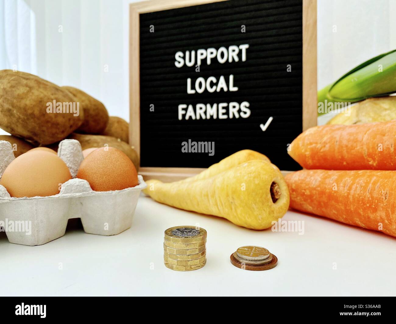 A selective focus view of a blurry farmer’s market sign behind fresh vegetables. Support local farmers. Fresh vegetables with British pound coins and loose change in foreground. - Smartphone Captured Stock Image
