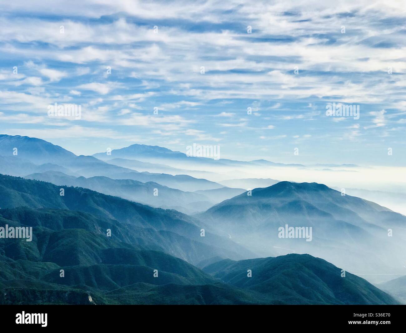 A scenic overview of layered mountains with a cloudy layer throughout under and cloud filled blue sky. Stock Photo