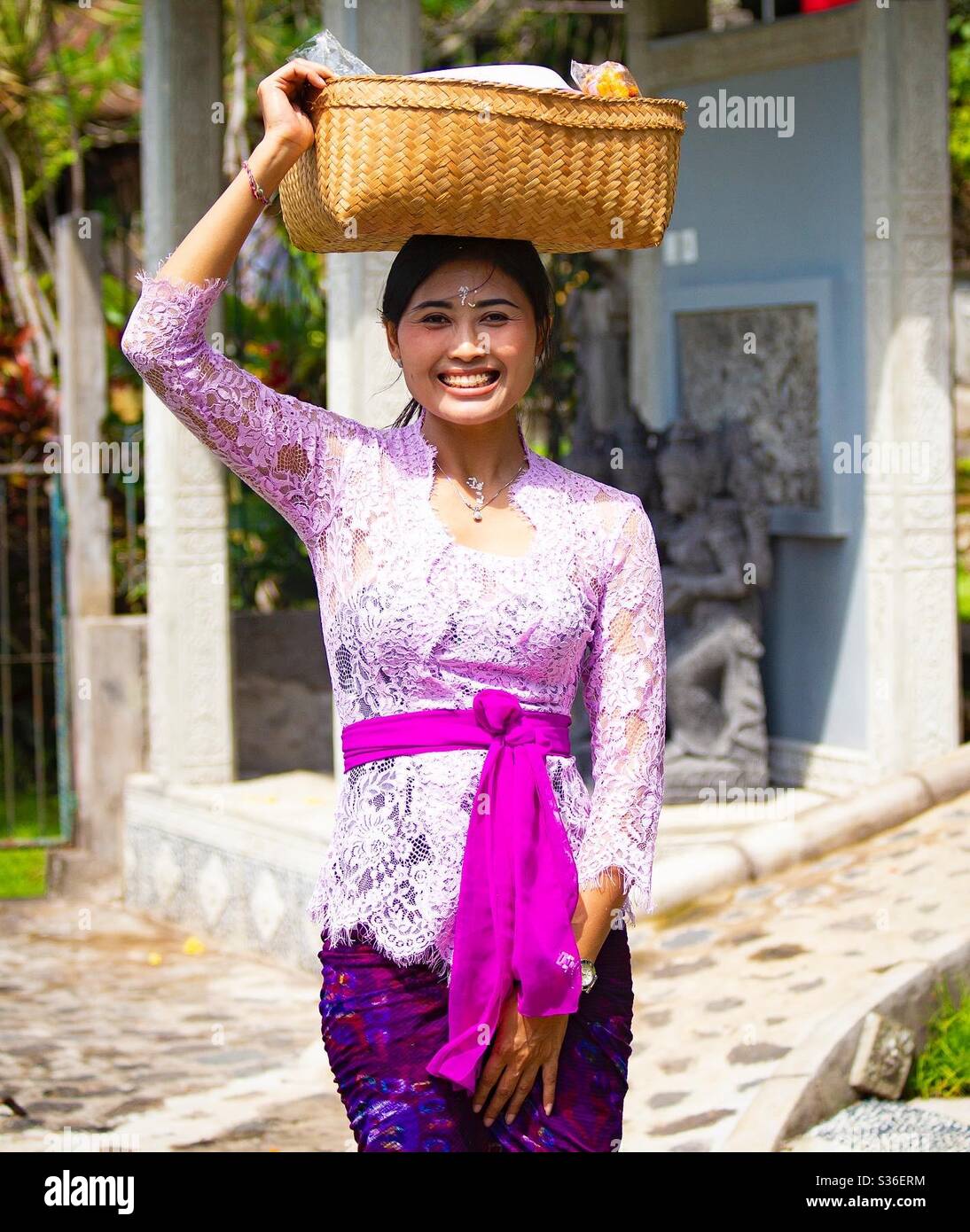 Beautiful smiling Balinese woman carrying a bucket with offering on her head, Bali, Indonesia - Smartphone Captured Stock Image