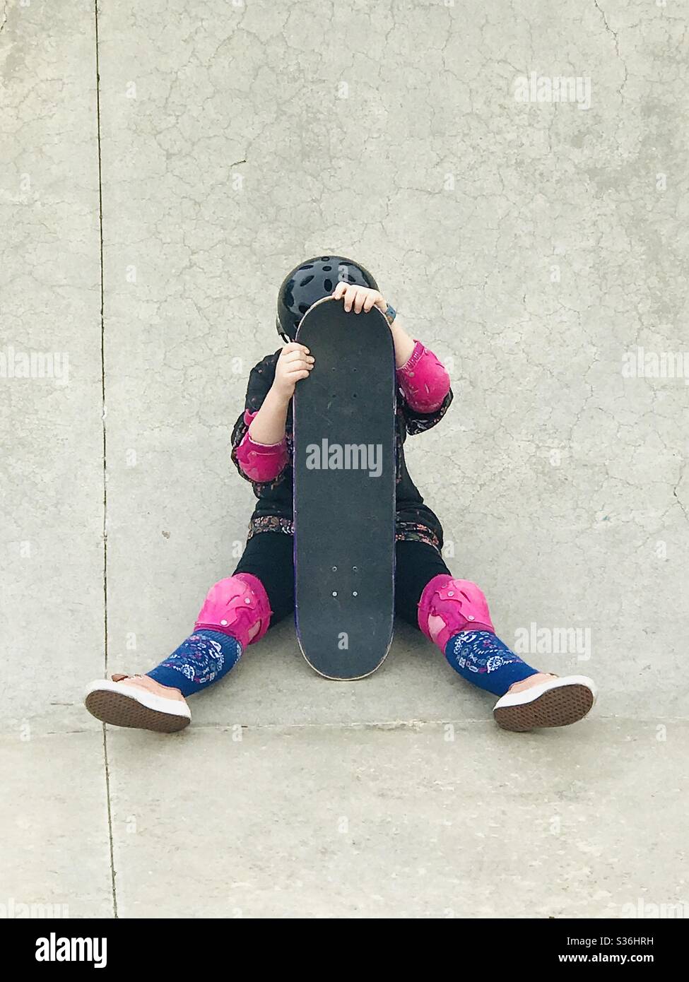 A girl sitting on the ground with a skateboard in from of her face. Stock Photo