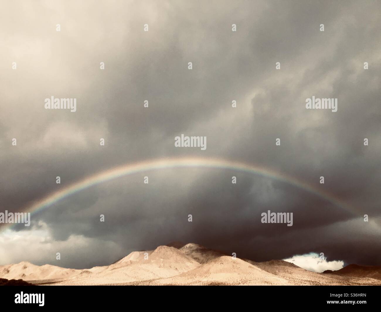 A full rainbow in an ominously dark and stormy sky over golden hills. Stock Photo