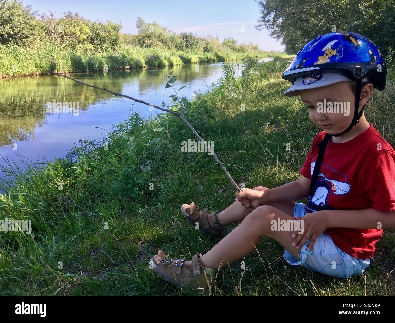 4 years old boy child sitting on lake shore holding a twig as fishing rod in summer, Ferto-to, Fertorakos, Hungary - Smartphone Captured Stock Image
