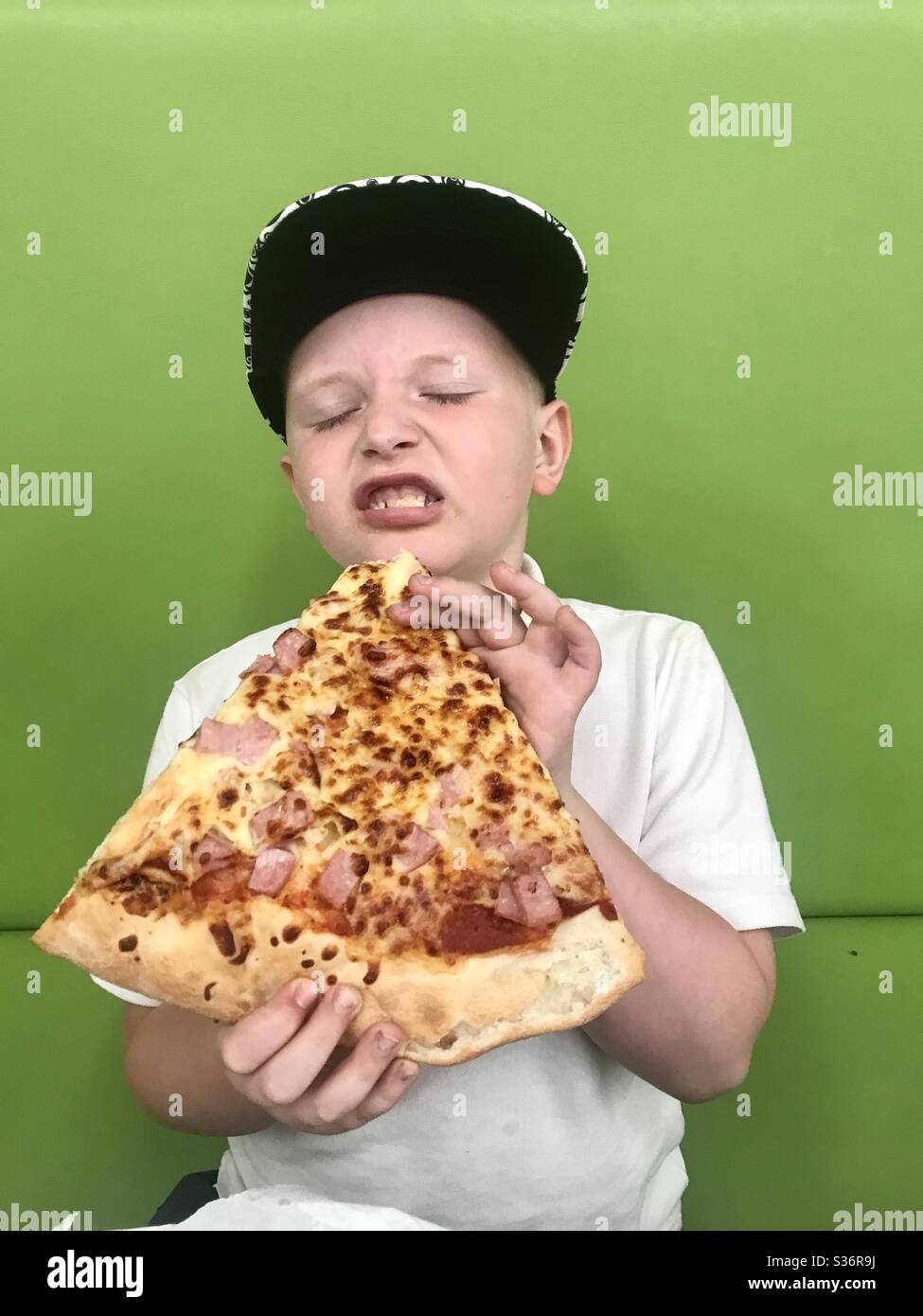 Young boy holds and eats a very large slice of pizza on a green background in school uniform Stock Photo