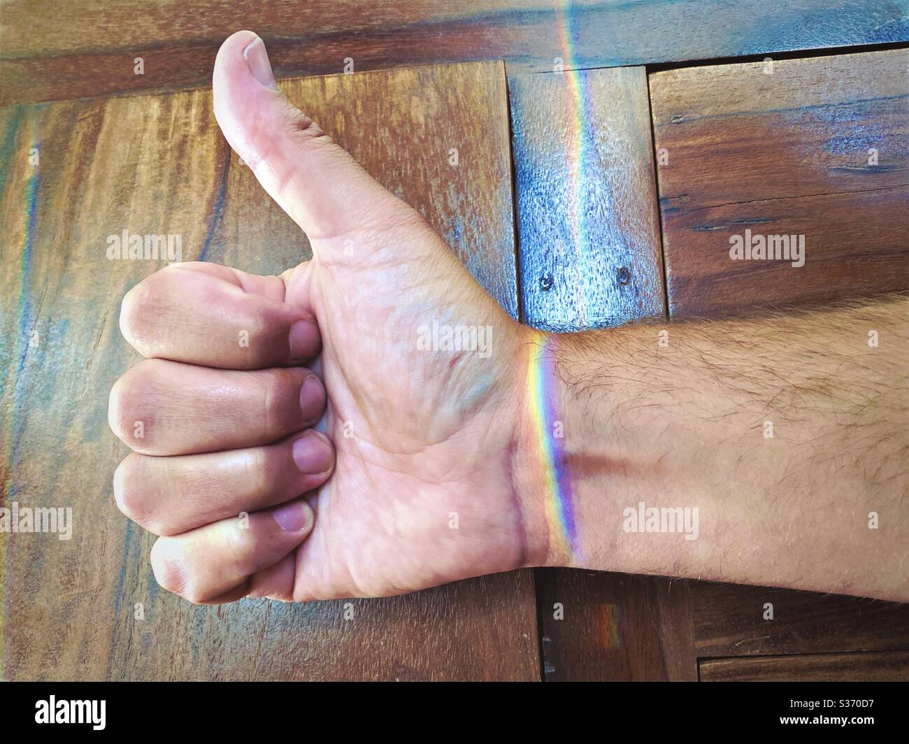 Happy LGBTQ Pride month! Close up view on the hand of a Caucasian male giving thumbs up gesture as light refracts off glass to create a prism colorful rainbow bracelet effect over the wrist - Smartphone Captured Stock Image