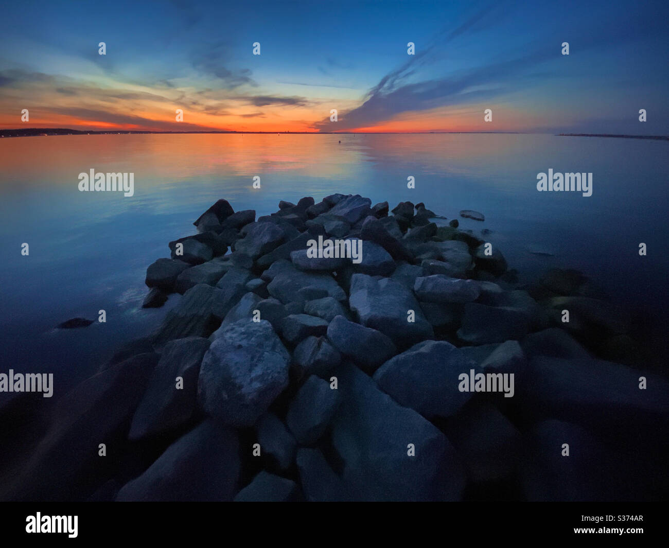 Long exposure shot with rocks in the Baltic Sea in foreground and dramatic sky with clouds and golden orange light at sunset in background, Germany - Smartphone Captured Stock Image