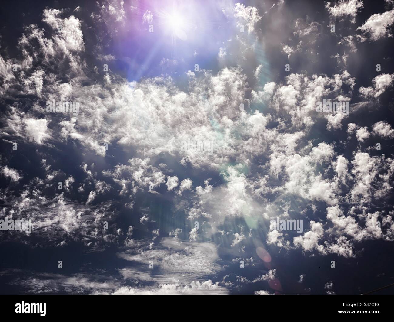 Abstract summer sky scene. White fluffy clouds are seen against a summery blue background with creative lens flare during midday sunshine. Bright light skyscape with copy space. - Smartphone Captured Stock Image