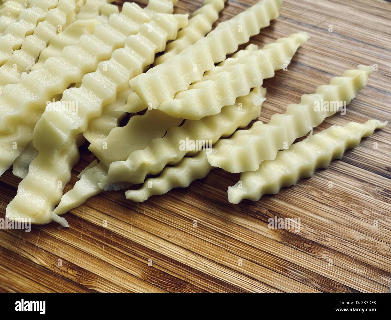 Preparing crinkle cut fries (US) or chips (UK) in the kitchen on a rustic wooden chopping board. Sliced potatoes with jagged edges ready for frying - Smartphone Captured Stock Image