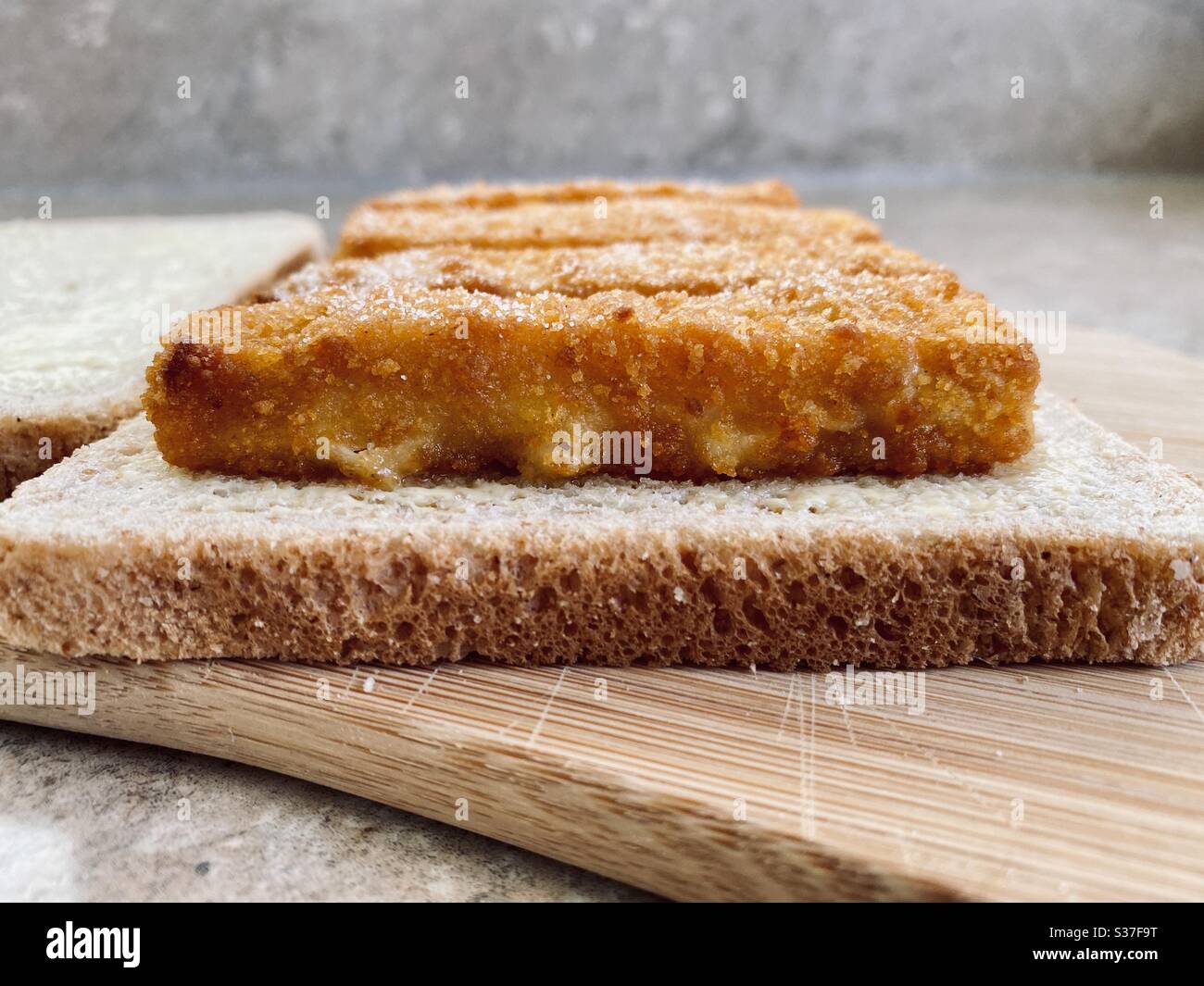 Closeup view of crispy fish fingers covered in salt and vinegar on buttered bread in the kitchen. Breaded fish finger sandwich preparation. - Smartphone Captured Stock Image