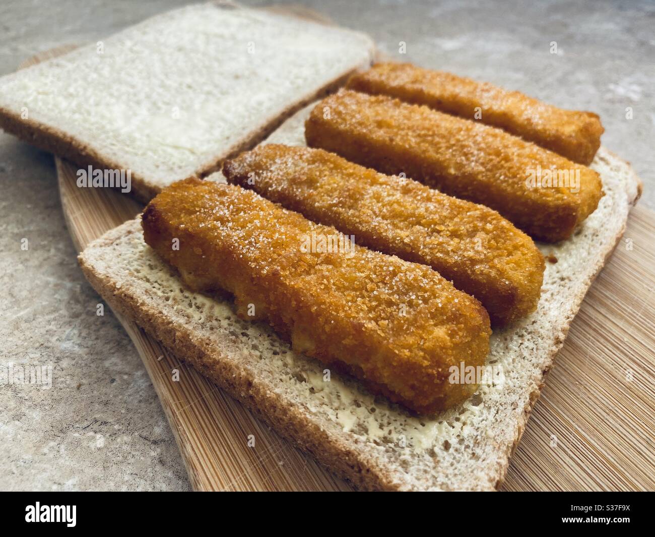 Closeup view of crispy fish fingers covered in salt and vinegar on buttered bread in the kitchen. Breaded fish finger sandwich preparation. - Smartphone Captured Stock Image