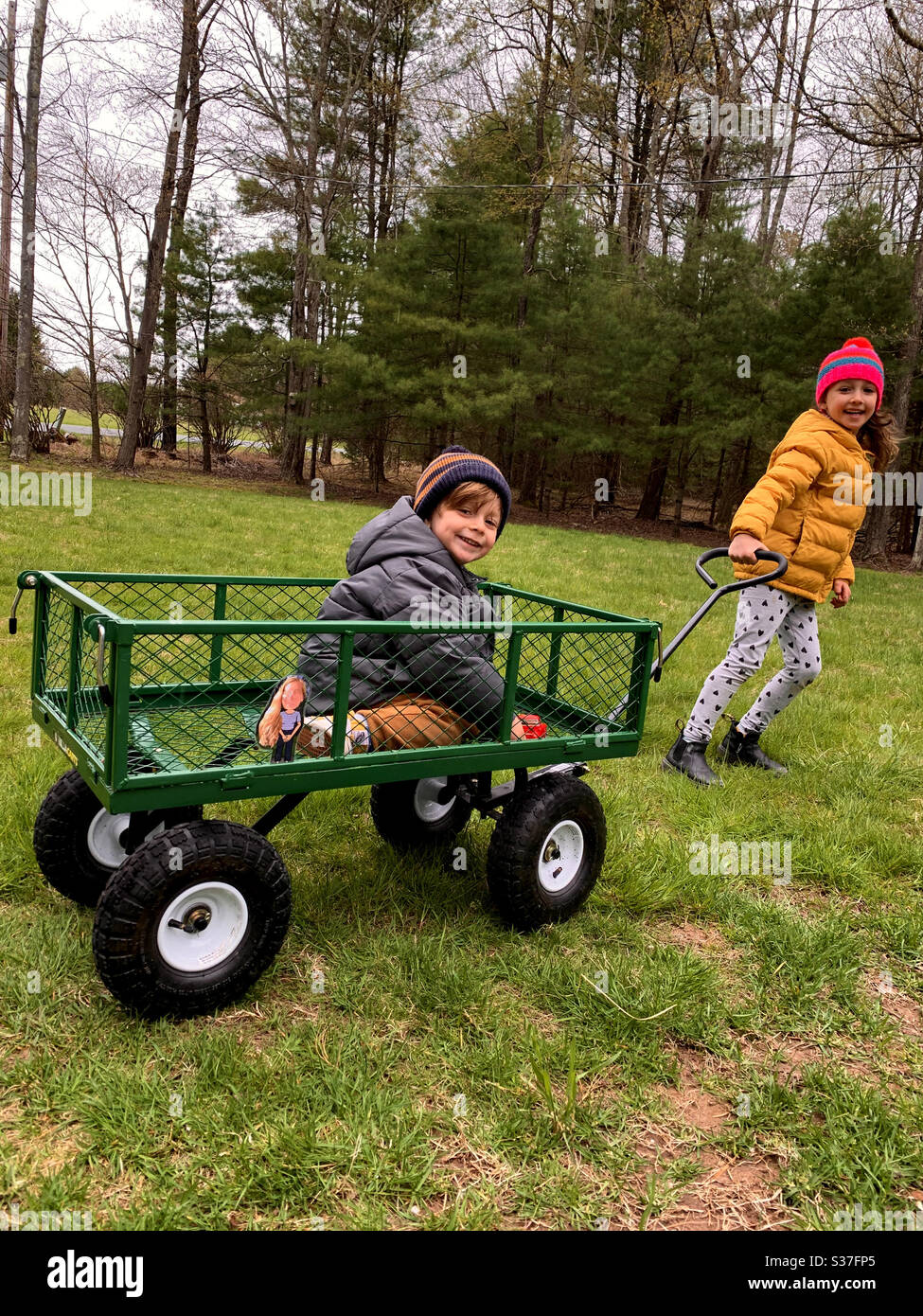 Happy older sister pulling her smiling toddler brother in a wagon dressed in warm jackets and hats. - Smartphone Captured Stock Image