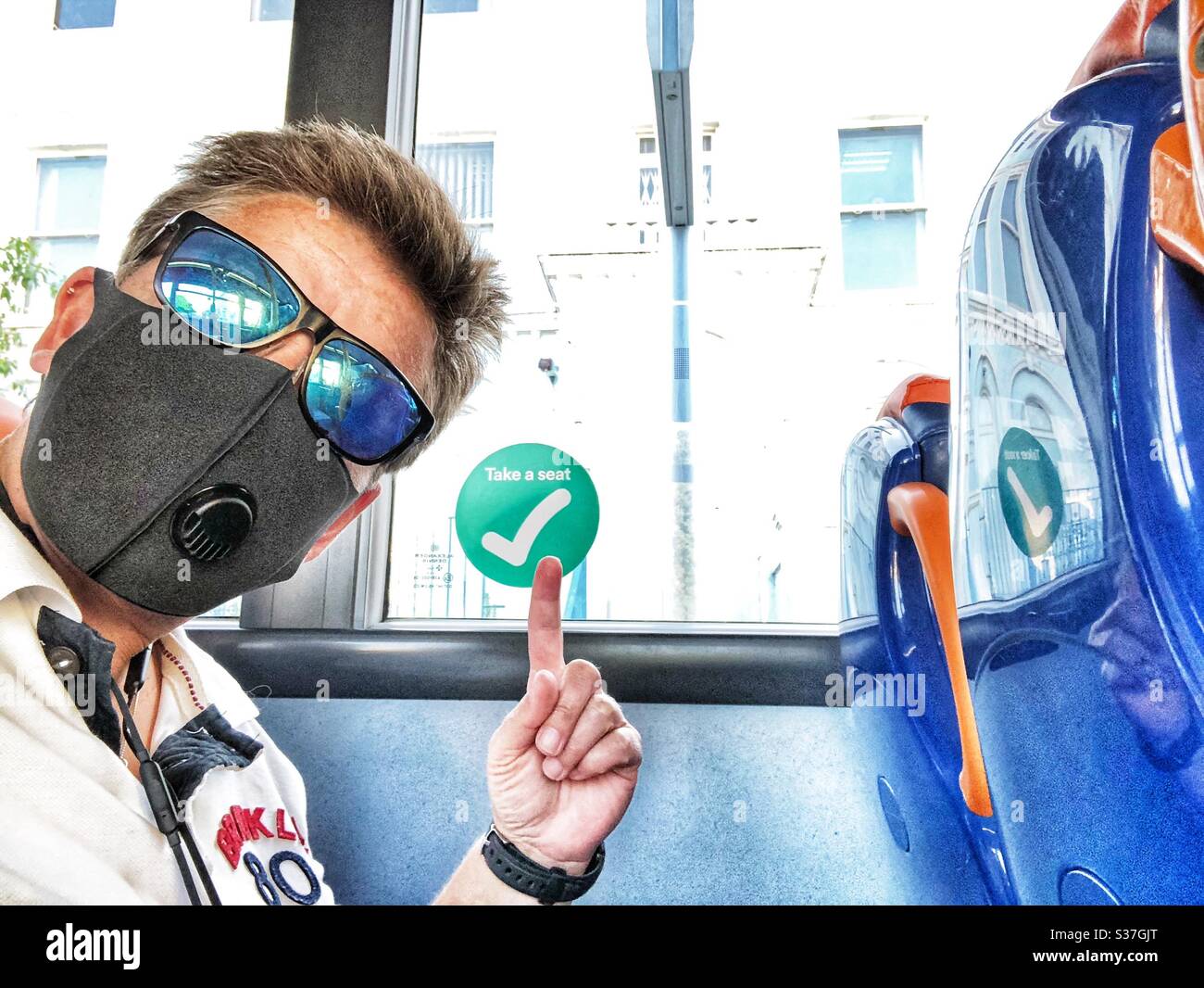 Woman on bus wearing face mask pointing at sign to indicate ok to sit on this seat - Smartphone Captured Stock Image