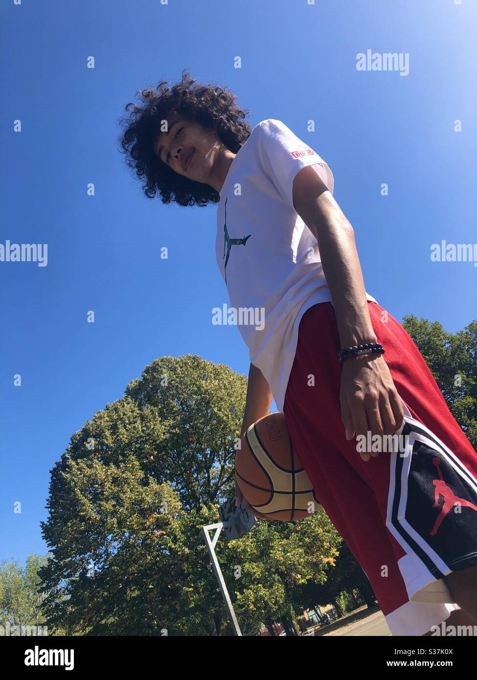 Young guy with curly hair looking down at the camera, getting ready to play basketball on a court in a London Park - Smartphone Captured Stock Image