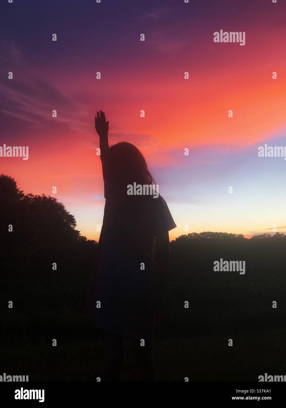 Silhouette of child with hand raised up against a vibrant summer sunset. - Smartphone Captured Stock Image