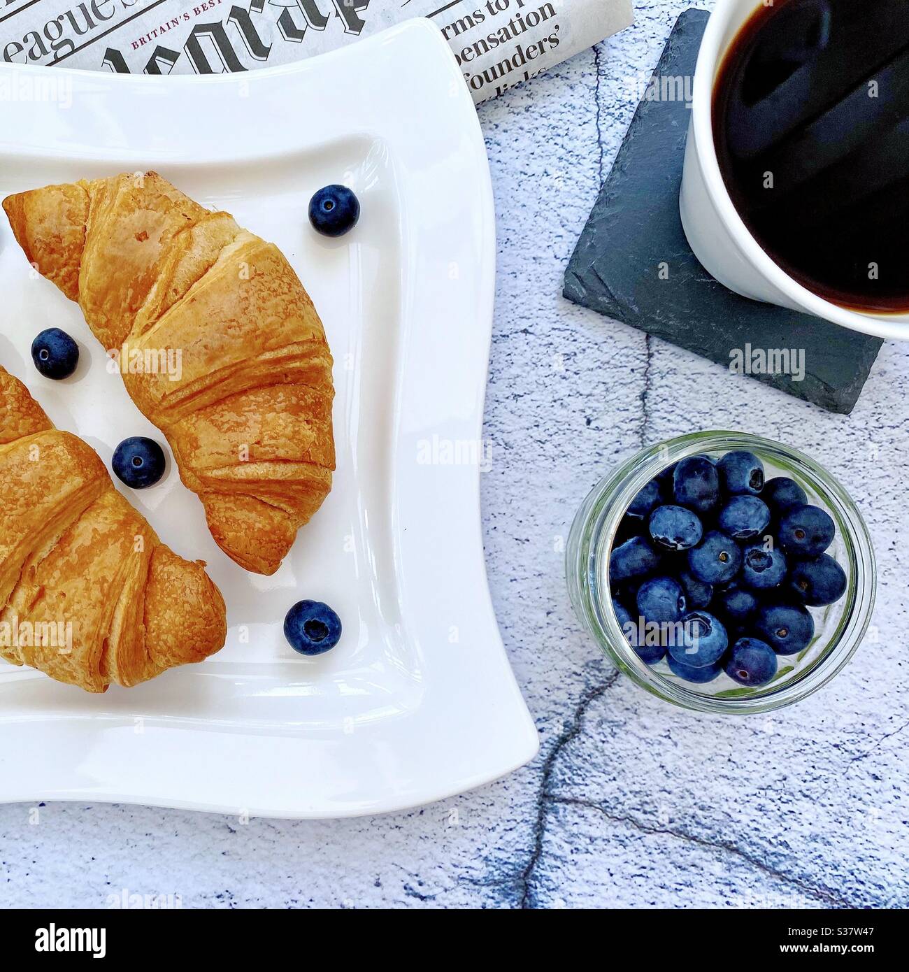 Closeup view of freshly baked croissants on a white plate with blueberries and grapes. Tasty breakfast with morning newspaper on a natural granite stone table. Served with coffee. - Smartphone Captured Stock Image