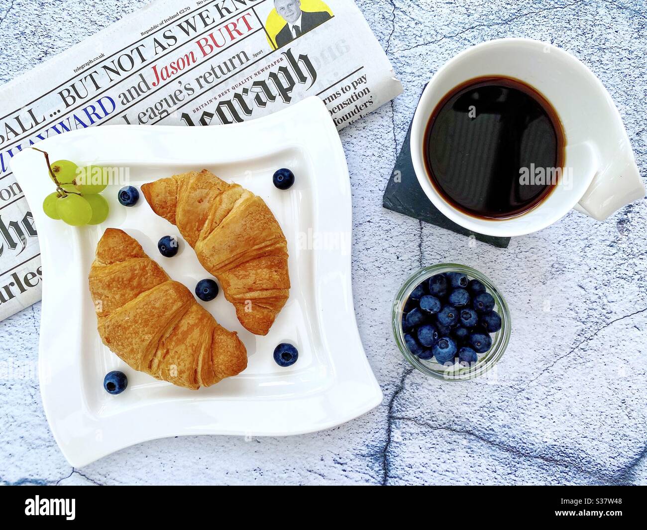Closeup view of freshly baked croissants on a white plate with blueberries and grapes. Tasty breakfast with morning newspaper on a natural granite stone table. Served with coffee. - Smartphone Captured Stock Image