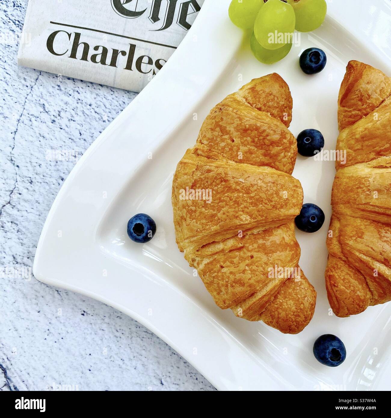 Closeup view of freshly baked croissants on a white plate with blueberries and grapes. Tasty breakfast with morning newspaper on a natural granite stone table. - Smartphone Captured Stock Image