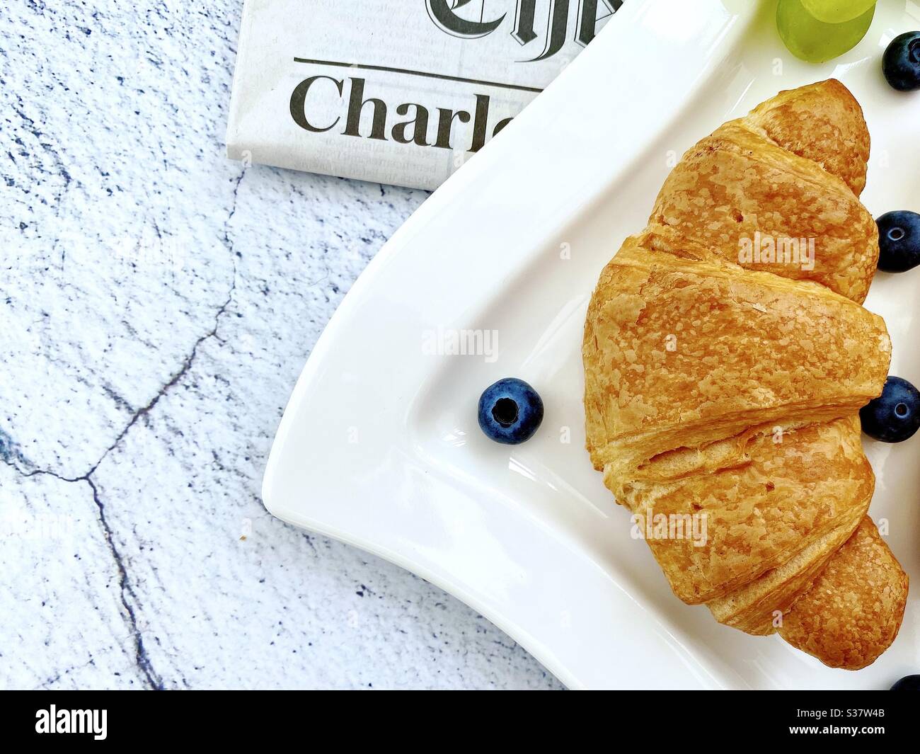 Closeup view of freshly baked croissants on a white plate with blueberries and grapes. Tasty breakfast with morning newspaper on a natural granite stone table. - Smartphone Captured Stock Image