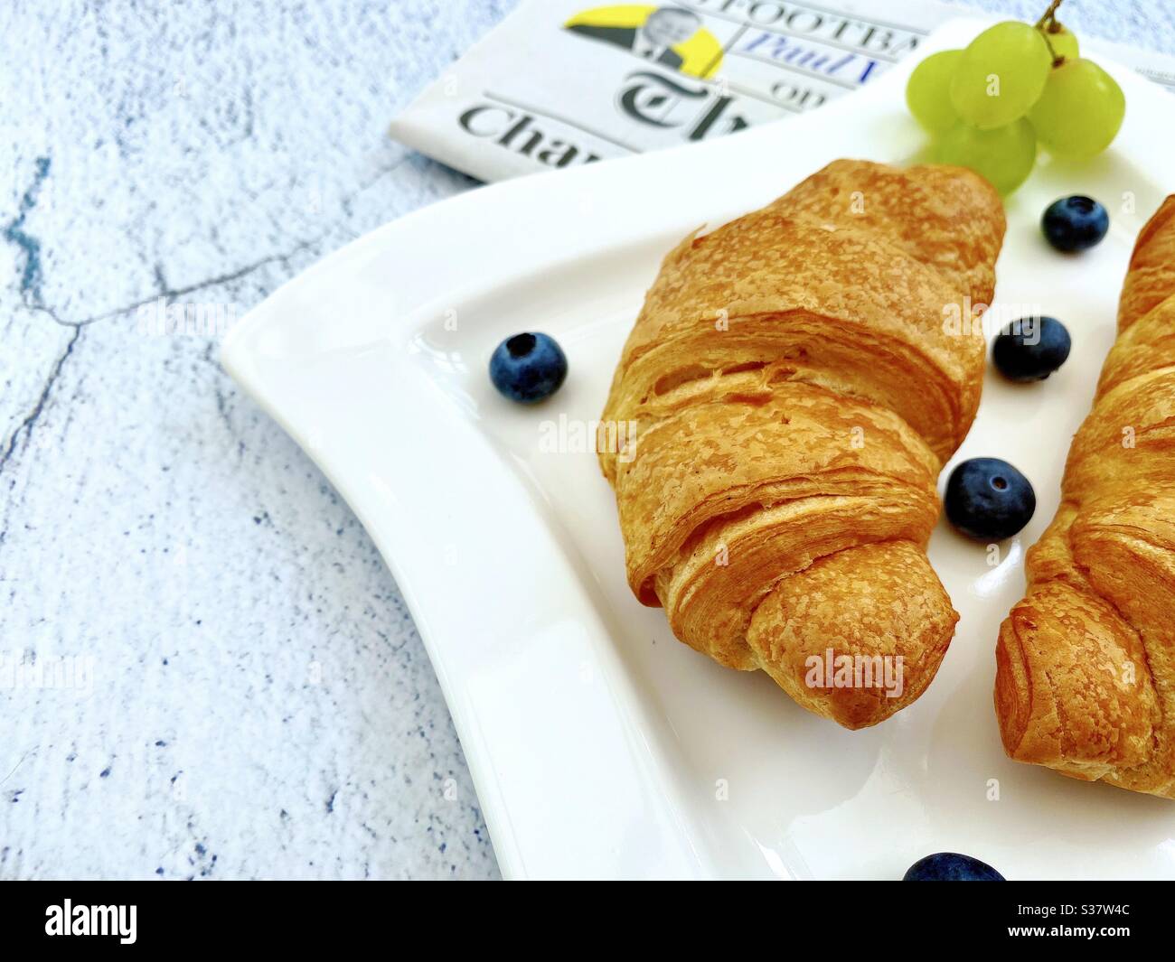 Closeup view of freshly baked croissants on a white plate with blueberries and grapes. Tasty breakfast with morning newspaper on a natural granite stone table. Stock Photo