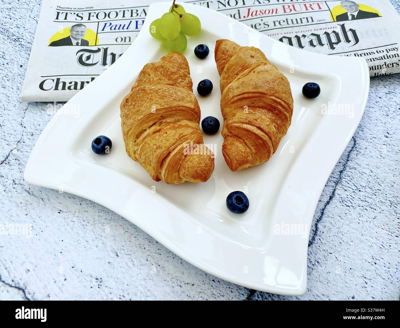 Closeup view of freshly baked croissants on a white plate with blueberries and grapes. Tasty breakfast with morning newspaper on a natural granite stone table. - Smartphone Captured Stock Image