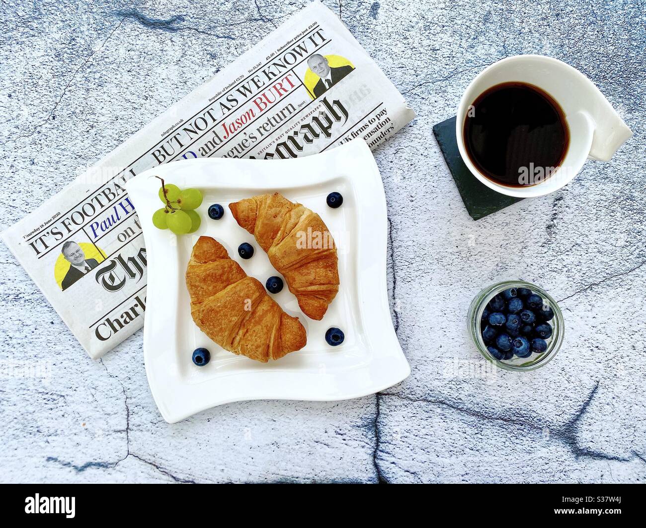 Closeup view of freshly baked croissants on a white plate with blueberries and grapes. Tasty breakfast with morning newspaper on a natural granite stone table. Served with coffee. - Smartphone Captured Stock Image