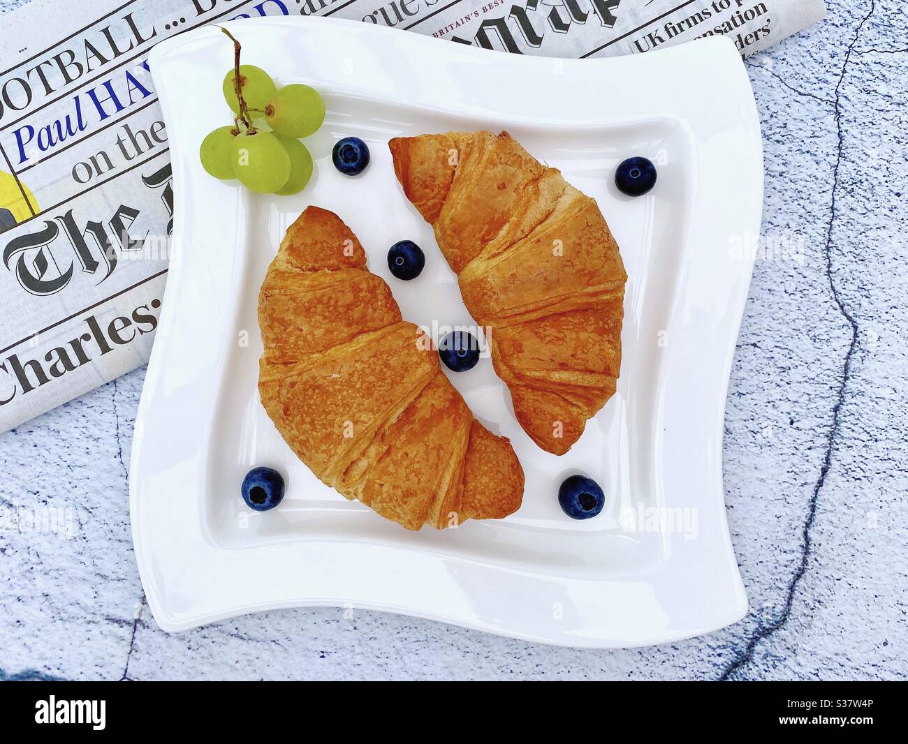 Closeup view of freshly baked croissants on a white plate with blueberries and grapes. Tasty breakfast with morning newspaper on a natural granite stone table. - Smartphone Captured Stock Image